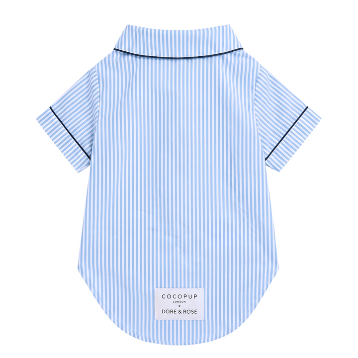 A light blue and white vertically striped short-sleeve dog shirt with thin blue piping along the collar and sleeve cuffs, featuring a white brand label at the bottom.