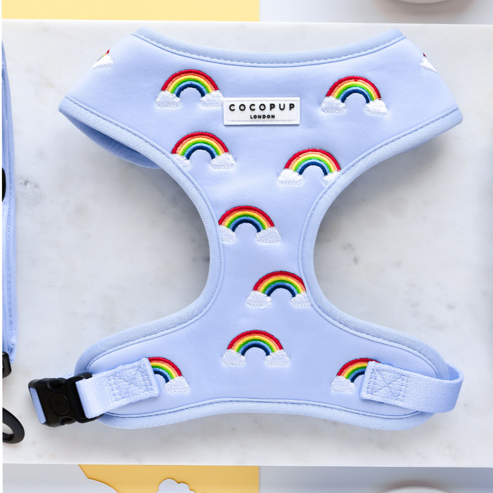 The Luxe Adjustable Neck Harness - Over The Rainbow, a pastel blue harness from Cocopup London, is embellished with an embroidered rainbow and cloud design. It features a black plastic buckle and has a label on the chest that reads "COCOPUP LONDON." The background is a white marble surface with a partially visible yellow object at the top.