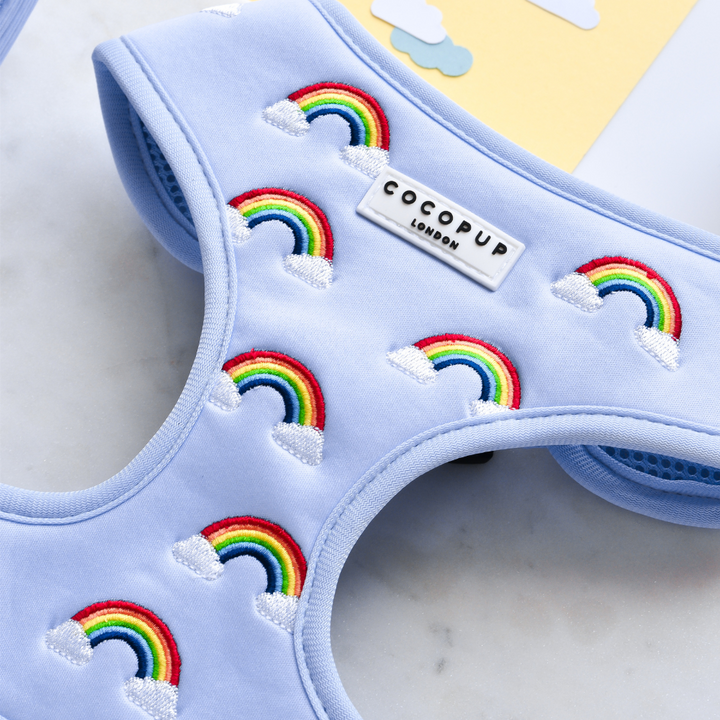 A close-up of the Luxe Adjustable Neck Harness - Over The Rainbow from Cocopup London showcases its light blue fabric adorned with embroidered rainbows and white clouds. A Cocopup London label is visible on the harness, which rests on a white marble surface with a beige envelope partially visible in the background.