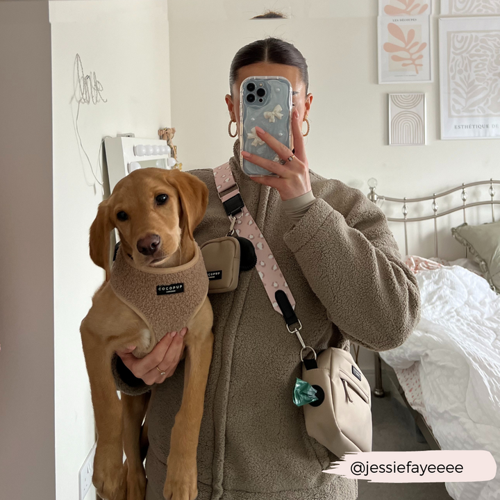 A person with their hair tied back is taking a mirror selfie while holding a small brown dog. The person is wearing a beige fleece jacket and crossbody bag adorned with the Cocopup London Bag Strap - Nude Chic Leopard, and the dog has on a beige harness. The room features a bed and wall art in the background. Instagram handle "@jessiefayeeee" is visible.