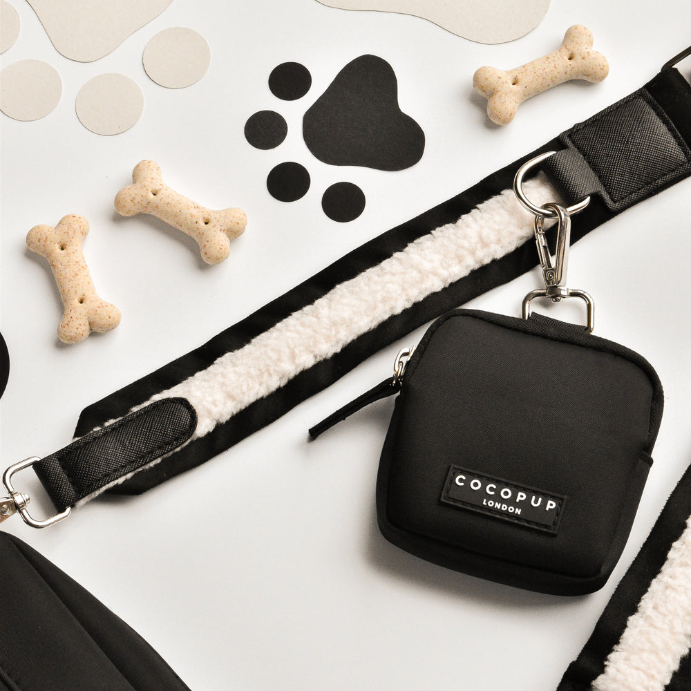 Image of various black and white dog accessories, including a black leash with a white fleece lining, a small Dog Walking Bag featuring the "Bag Strap - Black Aviator" from Cocopup London, bone-shaped treats, and black paw print cutouts scattered on a light background.