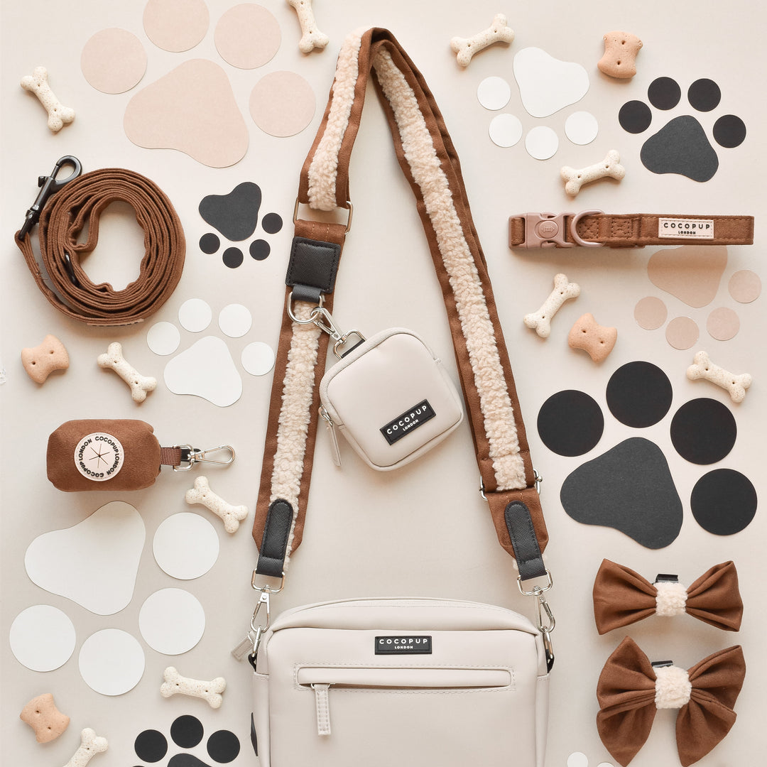 A flat lay image showcasing a variety of dog accessories, including a leash, harness, waste bag holder, bowtie, treats, and paw print cutouts. The items predominantly feature shades of brown, beige, and cream. A stylish Cocopup London Dog Walking Bag is neatly arranged against a light background with an eye-catching Brown Aviator Bag Strap.