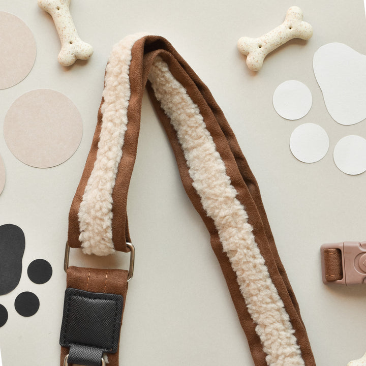 A brown and white fleece dog leash lies on a beige background. Surrounding it are assorted circular cutouts in various sizes and colors, a brown camera-shaped object, a Cocopup London Dog Walking Bag featuring the Brand Name's Bag Strap - Brown Aviator, and two bone-shaped dog treats. The composition has a playful and cozy aesthetic.