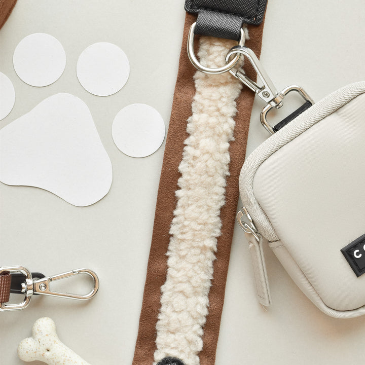 The image shows a white pet leash with a soft, fleece-like texture that features a metal clip. Next to the leash is a small, brown Cocopup London Bag Strap in an Aviator style with a black label and metal clasp. Decorative paw prints and a white bone-shaped toy are also visible.