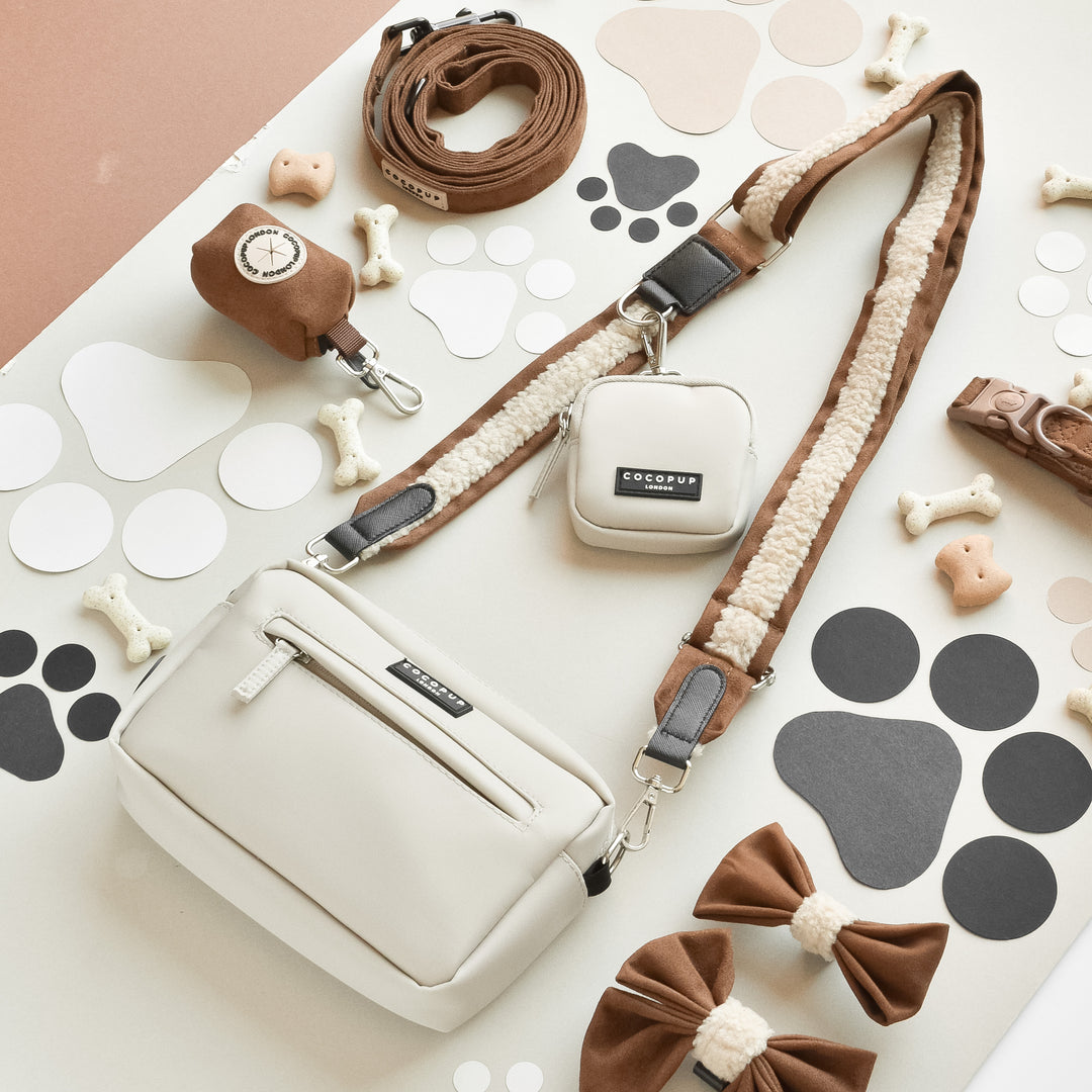 A stylish white and brown pet accessory set from Cocopup London is displayed, featuring a Brown Aviator Bag Strap, a smaller pouch, a leash, a waste bag holder, a bow tie, bone-shaped toys, and various paw print cutouts on a white surface.