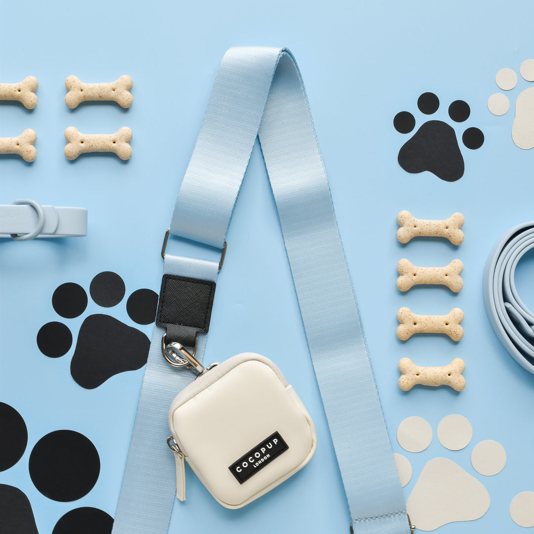 A light blue leash adorned with a white pouch, similar to the Cocopup London NAKD Bag Strap - Bubblegum, is laid out on a blue background. Nearby are tan bone-shaped dog treats and black and white paw print decals. The pouch features a black label with the text "COLOUP.