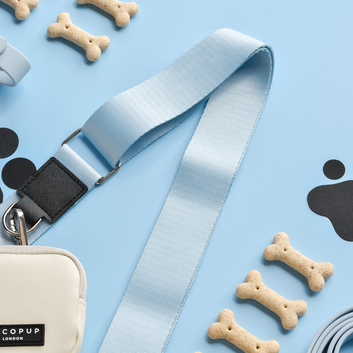 A dog leash featuring a pale blue Cocopup London NAKD Bag Strap - Bubblegum and a small white pouch, surrounded by bone-shaped dog biscuits and black paw print cutouts, arranged on a light blue background.