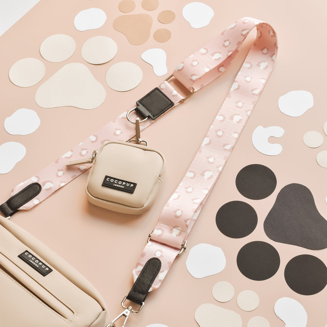 A beige handbag with black accents and a matching small pouch are placed on a pink and beige background adorned with various paw print patterns in black, white, and beige. The handbag, perfect as a Dog Walking Bag from Cocopup London, features a detachable strap named Nude Chic Leopard in light pink.