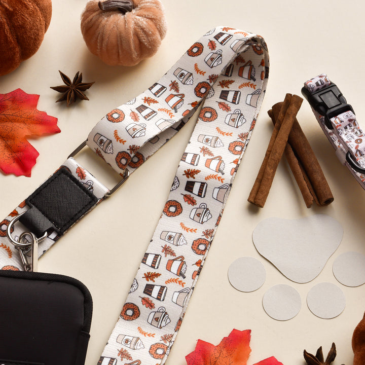 A fall-themed lanyard featuring autumn motifs like pumpkins, coffee cups, and leaves is surrounded by various seasonal items, including the Bag Strap - Autumn Puppuccino from Cocopup London, a black pouch perfect for an Autumn Puppuccino outing, cinnamon sticks, decorative pumpkins, maple leaves, star anise, and several circular cut-out shapes.