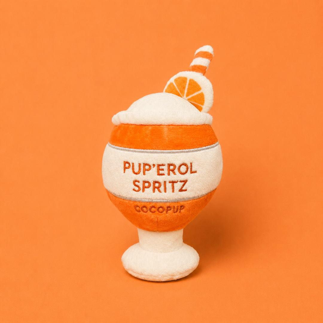 A small plush drinkware-shaped dog toy with the words 'PUP'EROL SPRITZ' and 'COCOPUP' on the front, shown on a solid orange background. 