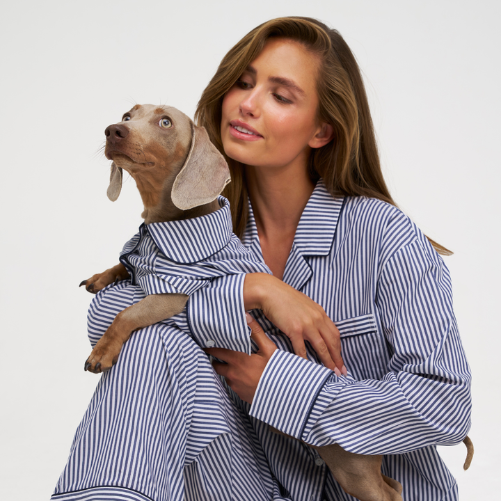A woman in blue and white striped pajamas holds a small dog wearing a matching pajama shirt against a plain light gray studio background.
