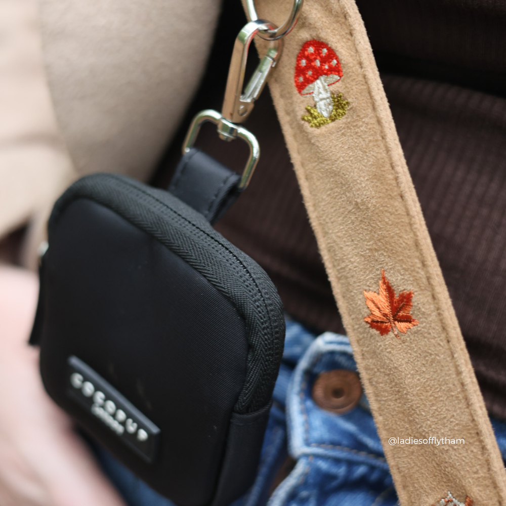 Close-up of a black fabric crossbody treat pouch with a tan suede strap featuring embroidered red and orange mushrooms and a leaf