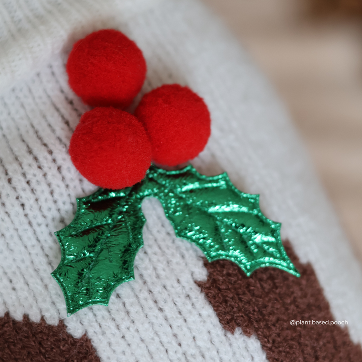 Close-up of a knitted holiday sweater featuring red felt pom-pom holly berries and shiny green felt holly leaves over a cream and brown knit pattern.